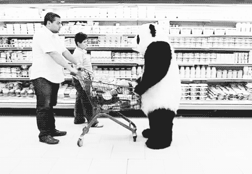 Person in a panda suite nocking over a cart of groceries that is being held by a man. They are in a grocery store and a younger person is standing next to the cart. Black and white.