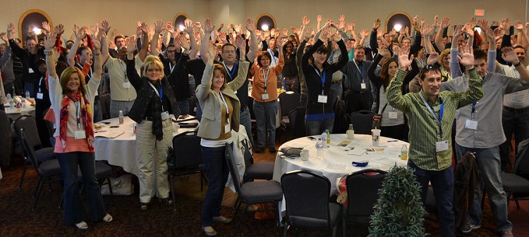 Kick off meeting with a crowd of people in a ballroom. They are standing at circular tables and holding their hands up in the air.
