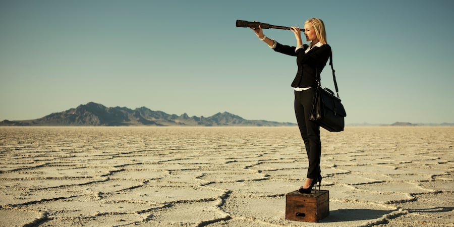 Person dressed in business clothes with a brief case in a desert standing on a wooden box looking through a monocular. 