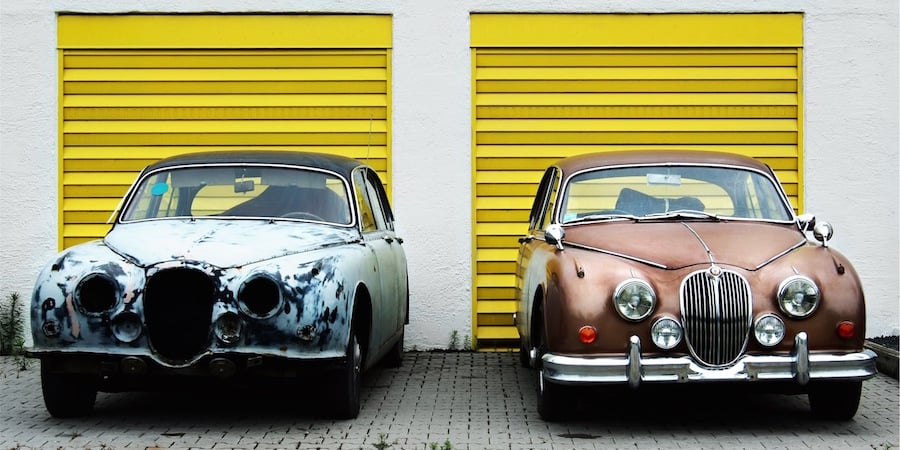 A old rusted car sitting next to the same type of car in restored condition. Behind them are two garage doors.