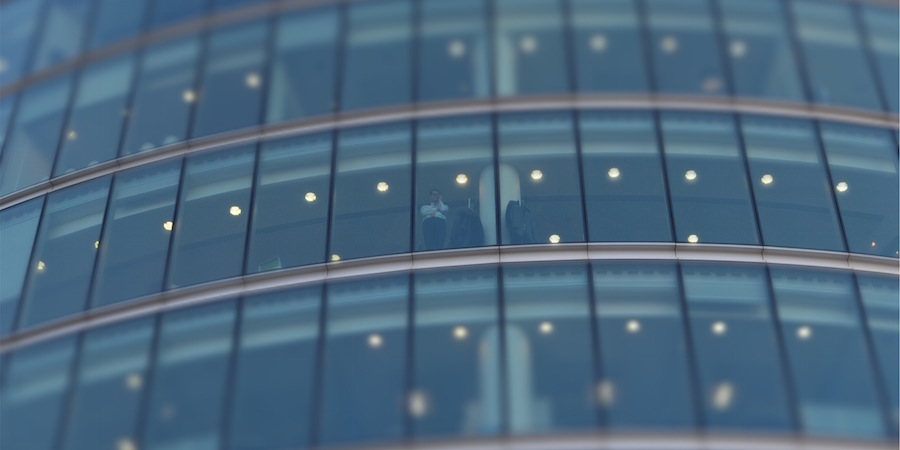Building exterior made of full height windows. Inside i n a business person ins a suite talking on a phone.