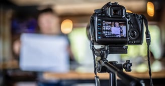 DSLR camera on a tripod pointed at a person holding a chart. The person holding the chart is blurry in the background but clear in the camera display.