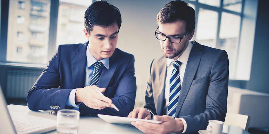 Two business people sitting at a table looking at a tablet. They are wearing suites and ties and sitting in front of windows. On the table is a laptop, coffee cup, and water glass.