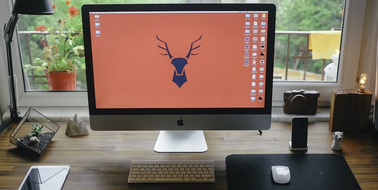 Apple iMac desktop on a desk in front of a window. The desktop image is an icon of a deer in the center.