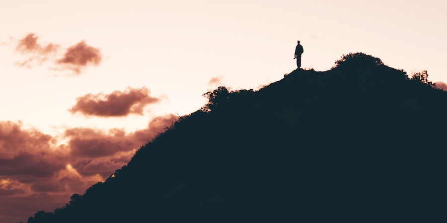 Person in silhouette standing on top of a hill with clouds in the background.
