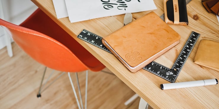 Notebook, corner ruler, marker, and other items sitting on a wooden desk.