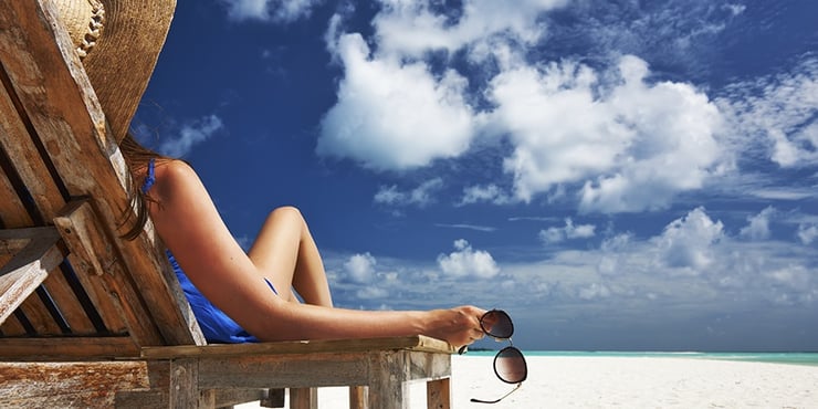 Person sitting on a reclined wooden chair on a beach looking at a blue sky with white clouds.
