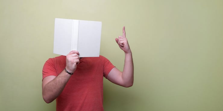 Person standing in front of a blank wall holding a book up to cover their face. Their left hand is pointing up with their index finger.