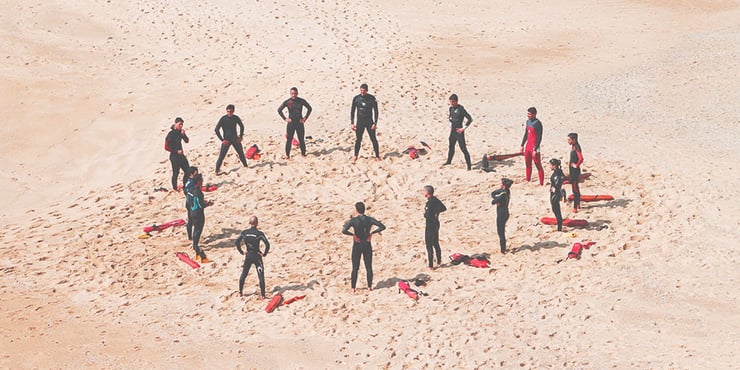 Life guards in wet suites standing in a circle on a beach with rescue floaters.