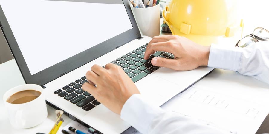 Hands typing on a laptop. There is a copy mug and tape measure on the left and glasses, mug full of pencils, and hard hat to the right.