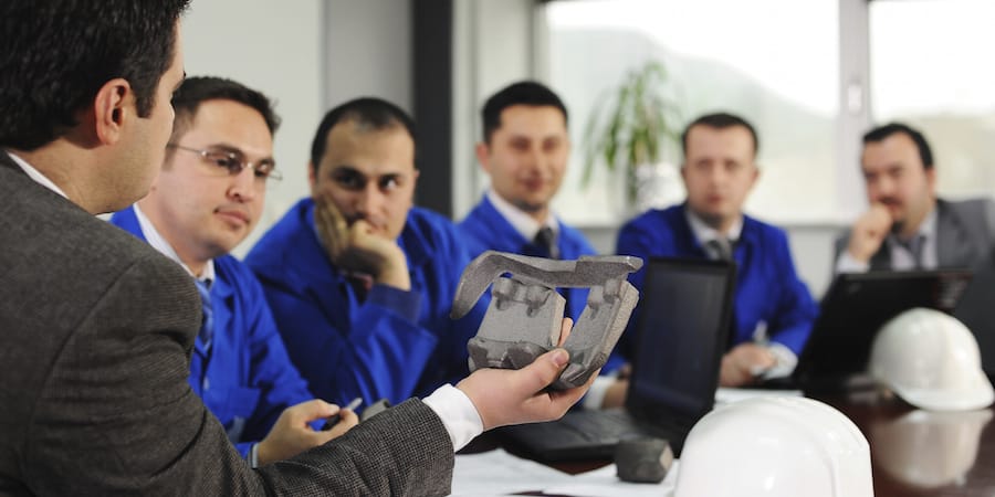 Manufacturing team sitting at a conference room table. Person in foreground is holding a casted part. There are laptops and hard hats on the table.
