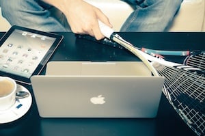 A person holding a tennis raquet looking at a MacBook and iPan that are sitting on a desk.