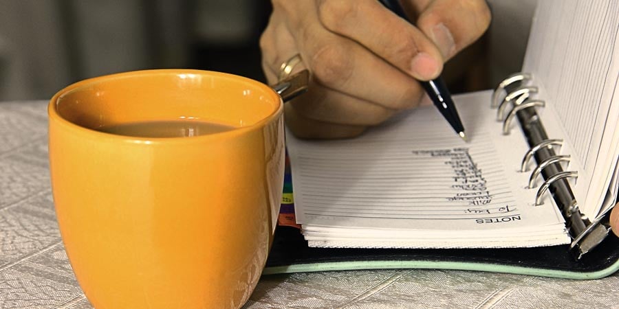 Mug on a table with a notebook. Someones hand is writing down to dos in the notebook.