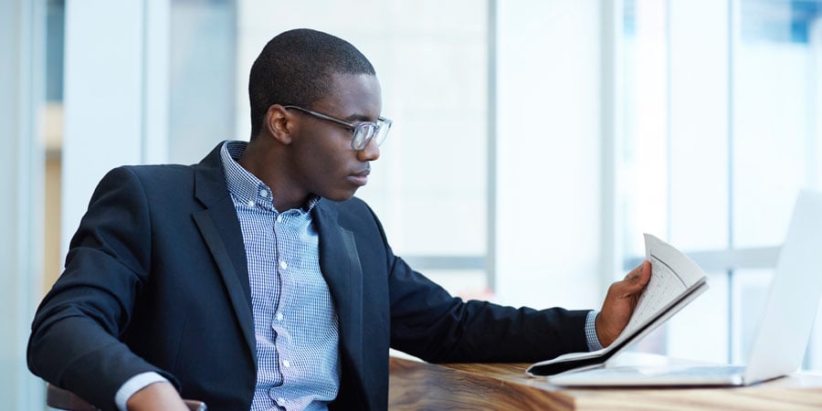 Person sitting at a carved wood desk reading a paper. There is also a laptop on the desk.