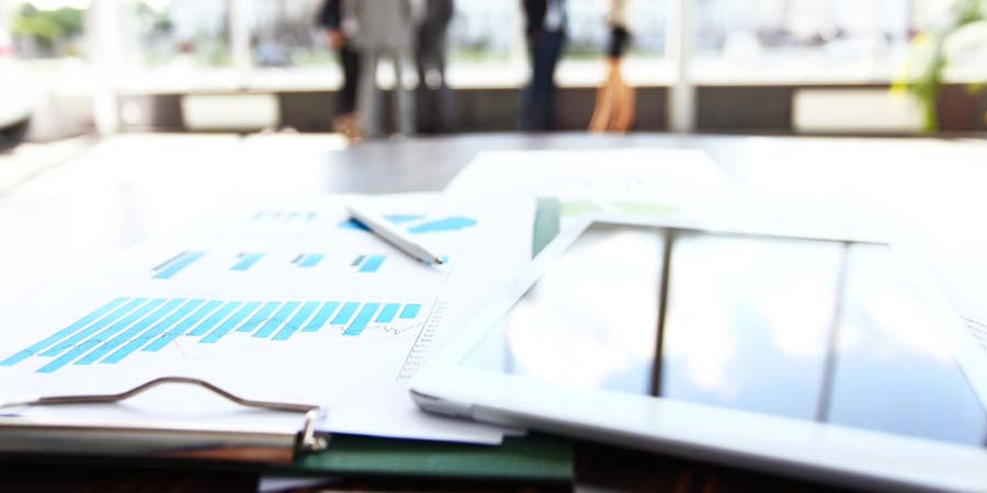 Tablet and clipboard on table with office staff in a blurred background. On the clipboard are graphs and a pen.