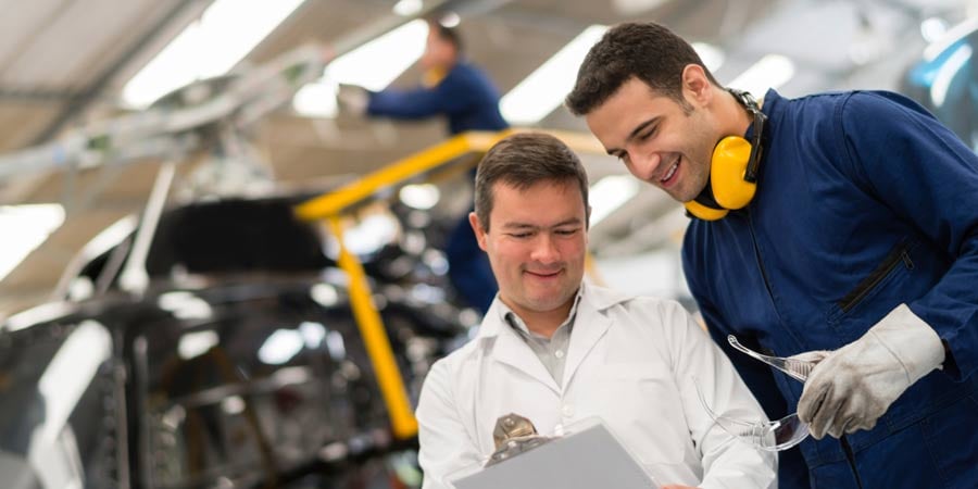 Two people in a helicopter manufacturing plant looking at a clipboard. One has noise protection earmuffs around their neck and is holding safety glasses.