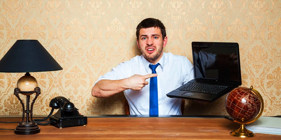 Person looking annoyed holding a laptop and pointing at it. He is in front of a desk that has a lamp, rotary phone, and globe on it.