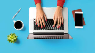 Hands typing on a laptop on a blue surface along with a smart phone, coffee, plant, and pen.