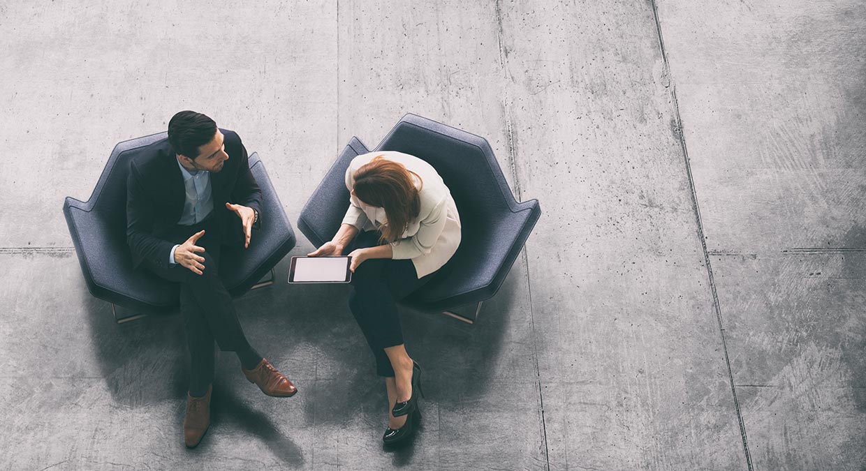 Overhead shot of two people talking in chairs. One is holding an tablet computer. The floor is concrete.