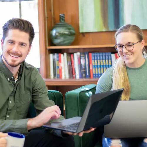 Two Weidert Group employees. One is holding a laptop. They are sitting in front of books and art.